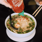 hand pouring sriracha on a bowl of pho