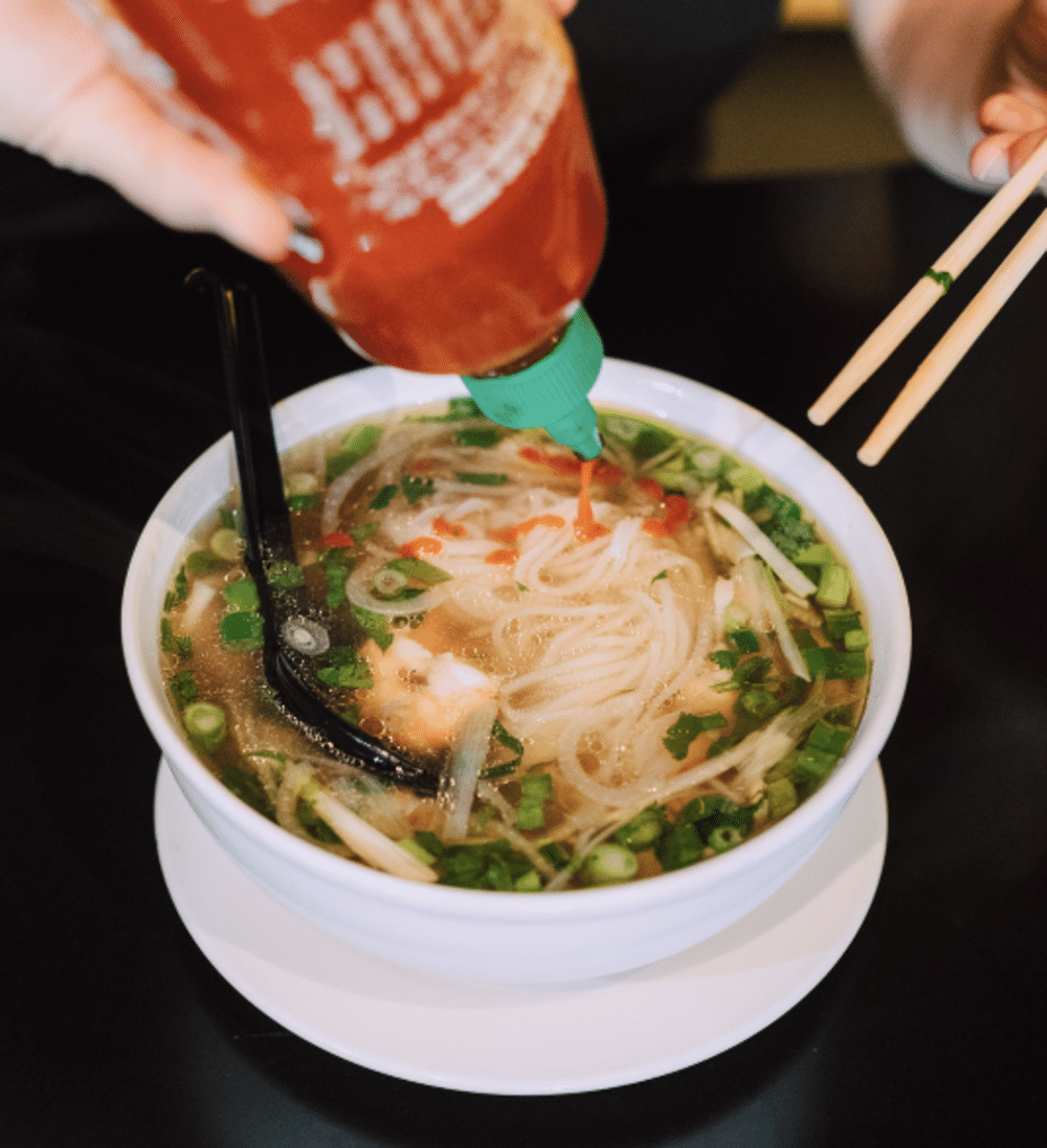 hand pouring sriracha on a bowl of pho