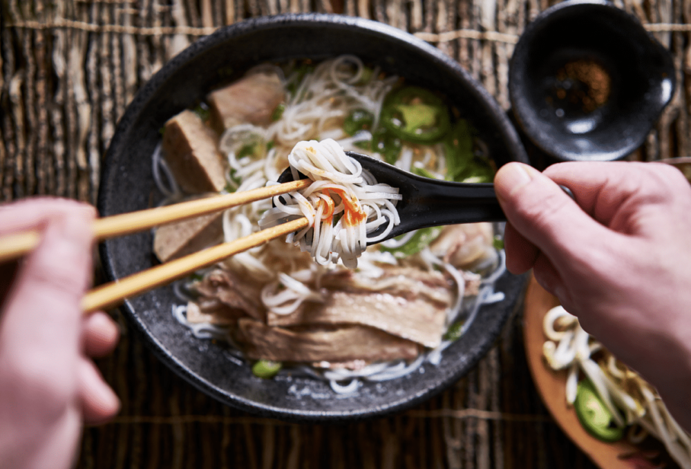 hands using a Chirenge and chop sticks over a bowl of pho